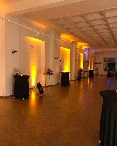 Ein geräumiger, eleganter Raum mit Holzböden, weißen Wänden und warmer gelb-oranger Beleuchtung - perfekt für diejenigen, die eine Weihnachtsfeier planen oder eine DJ-Firma Weihnachtsfeier Österreich ausrichten möchten. Hohe, runde Cocktailtische sorgen für eine einladende Veranstaltungsatmosphäre.