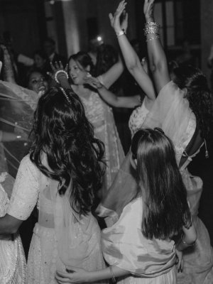 Eine Gruppe von Frauen in traditionellen Kleidern tanzt freudig mit erhobenen Armen bei einer Indoor-Veranstaltung, während andere im Hintergrund zusehen. Das Bild ist in schwarz-weiß gehalten.