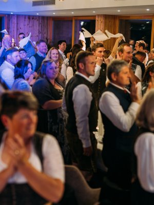 Eine große Gruppe traditionell gekleideter Menschen steht und klatscht bei einer festlichen Veranstaltung in einer Halle, viele lächeln und schwenken weiße Servietten in der Luft. Die Atmosphäre wirkt fröhlich und feierlich.