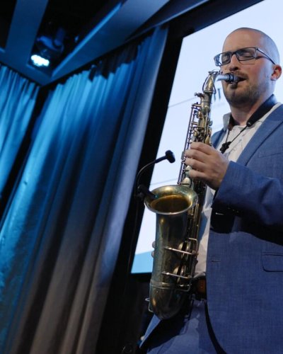 Ein Mann in einem blauen Anzug spielt auf der Bühne ein Saxophon. Hinter ihm befinden sich ein blau beleuchteter Hintergrund und ein Bildschirm mit verschiedenen Logos.