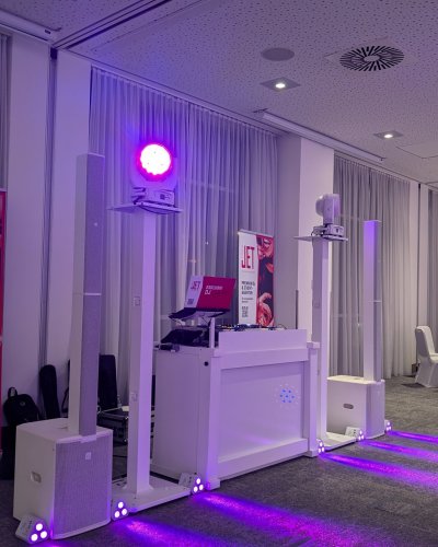 Ein professionelles DJ-Pult mit Lautsprechern, Lichtern und elektronischem Equipment erstrahlt unter violettem Licht in einem modernen Raum mit großen Fenstern, weißen Vorhängen und Werbebannern im Hintergrund.