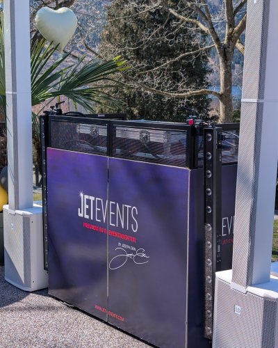 Ein DJ-Pult mit einem "JET EVENTS"-Banner steht an einem sonnigen Tag im Freien, flankiert von zwei Lautsprechersäulen. Ein großer weißer Herzballon hängt in der Nähe, und im Hintergrund sind Bäume und grünes Gras zu sehen.