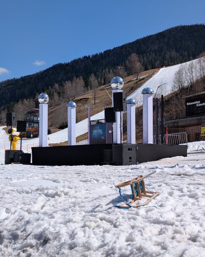 Eine kleine Bühne mit hohen Säulen, die von reflektierenden Kugeln gekrönt werden, steht auf Schnee, umgeben von Lautsprechern. Im Vordergrund liegt ein Holzschlitten auf dem Schnee. Hinter der Bühne befinden sich Hügel, Bäume und eine Skipiste.