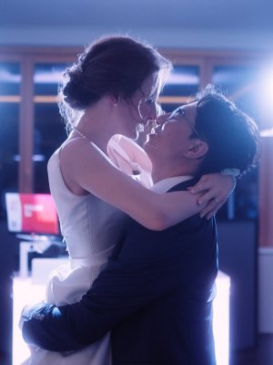 Ein formell gekleidetes Paar genießt einen intimen Moment, umarmt sich und lächelt einander auf einer beleuchteten Tanzfläche während einer Hochzeitsparty an, mit hellen Lichtern und verschwommenen Figuren hinter ihnen - perfekt für einen Hochzeits-DJ.