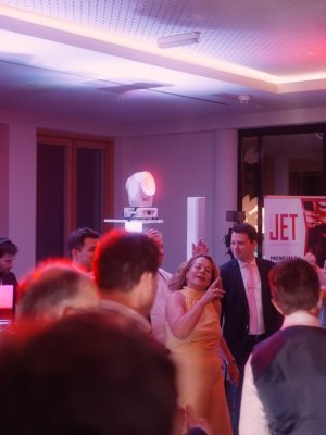 Eine lebhafte Indoor-Hochzeitsparty mit tanzenden und geselligen Menschen unter roter und lila Beleuchtung. DJ-Equipment, ein "JET"-Schild und ein professioneller Hochzeits-DJ sorgen für die festliche Stimmung, während die formell gekleideten Gäste die Feier genießen.