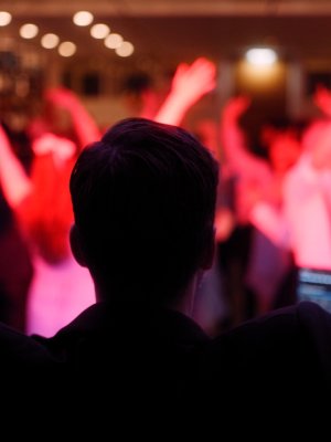 Eine Person mit erhobenen Armen steht vor einer lebhaften Menge, die unter bunten Lichtern in einem schwach beleuchteten Raum tanzt und die festliche Stimmung einer Feier einfängt - die perfekte Inspiration für Hochzeitsmusik oder jeden, der einen DJ für Hochzeiten sucht.