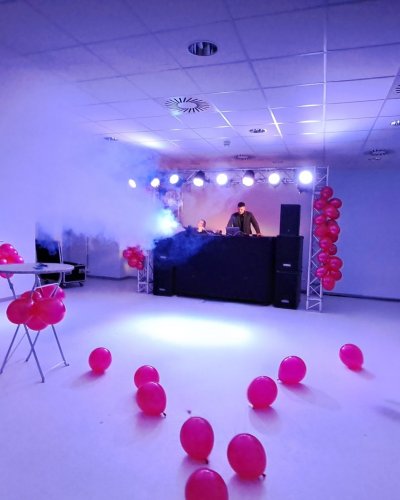 Ein weitgehend leerer Partyraum mit roten Luftballons, die auf dem Boden verstreut und an Tischen befestigt sind. Zwei Event-DJs einer Top-DJ-Agentur stehen hinter einem Pult, bunte Bühnenlichter und Raucheffekte sorgen für eine aufregende Stimmung vor modernen Decken und weißen Wänden.