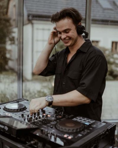 Ein lächelnder DJ mit Kopfhörern steht hinter einem Mischpult und bedient die Regler mit einer Hand. Er befindet sich in einem Raum mit Glaswänden und einem Blick ins Freie im Hintergrund.