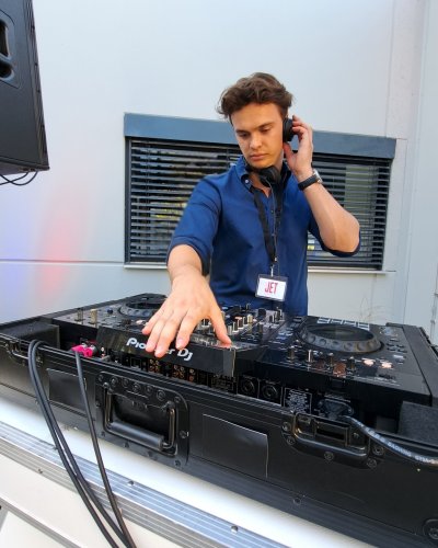 Ein DJ, der ein Namensschild und Kopfhörer trägt, arbeitet im Freien mit einem Mischpult, wobei er eine Hand an den Reglern hat und die andere eine Ohrmuschel hält. Im Hintergrund sind Fenster und Gebäudeteile zu sehen.