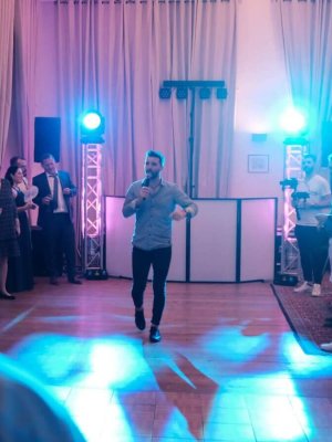 Ein Mann singt auf einer Tanzfläche in ein Mikrofon, umgeben von einer Menschenmenge. Blaue und violette Lichter erzeugen eine energiegeladene Atmosphäre, während die Leute zuschauen. Die Kulisse scheint eine formelle Veranstaltung oder Feier in einem geräumigen Raum mit Vorhängen zu sein.