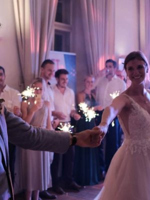 Ein frisch verheiratetes Paar tanzt fröhlich und hält sich an den Händen bei seinem Empfang. Die Braut im weißen Kleid und der Bräutigam im grauen Anzug sind in einem schwach beleuchteten Raum von Gästen mit Wunderkerzen in der Hand umgeben.