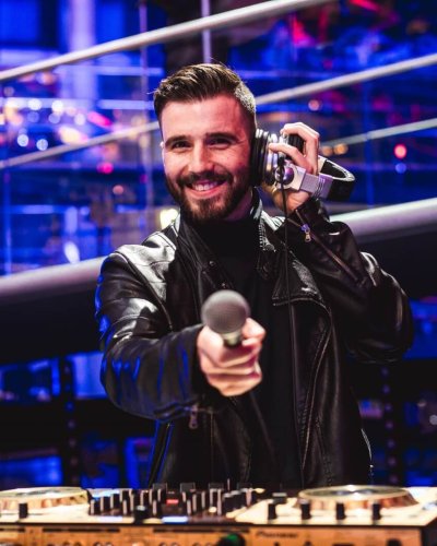 Ein DJ für Weihanchtsfeiern steht an seinem DJ Pult in einer in blau geflutetet Eventhalle. Mit seinen Kopfhörern am Ohr deutet er mit seinem Mikrofon in die Kamera.