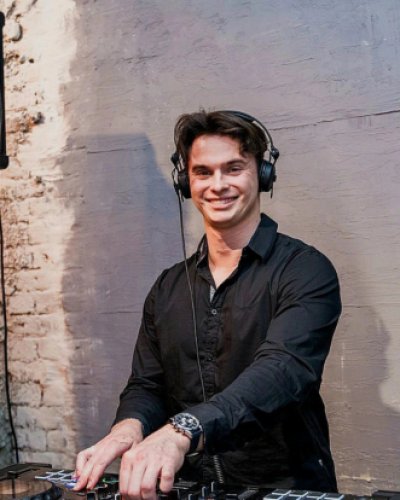 Eine Person, die Kopfhörer und ein schwarzes Hemd trägt, lächelt, während sie eine DJ-Anlage vor einer strukturierten Ziegel- und Gipswand benutzt.