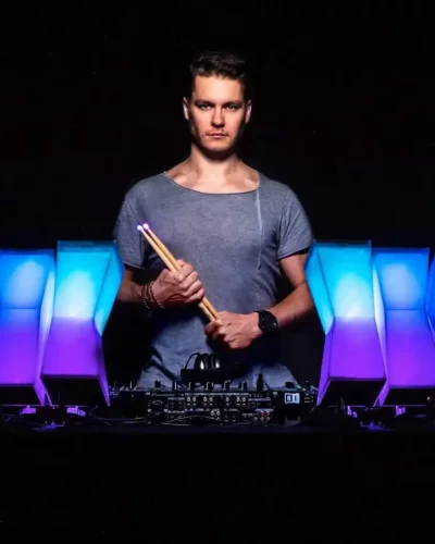 Ein Mann in einem grauen Hemd steht mit Drumsticks in der Hand vor einem DJ-Equipment. Hinter ihm erleuchten bunte Lichter in Blau- und Lilatönen segmentierte Panels in einer dunklen Umgebung.