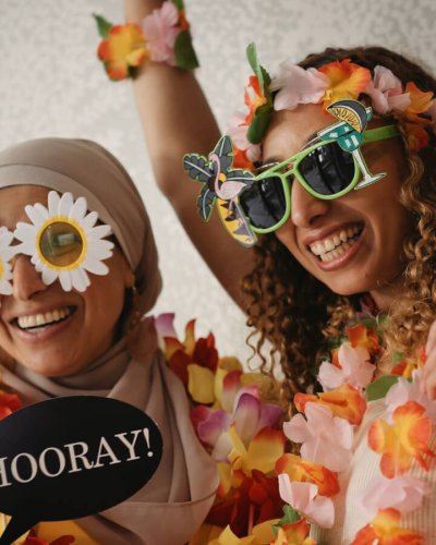 Zwei Frauen, eine davon trägt einen Hijab, feiern auf einer Party mit bunten Blumenketten, neuartigen Sonnenbrillen und einem "Hurra!"-Schild vor einem Hintergrund mit Vintage-Muster und einem Boom