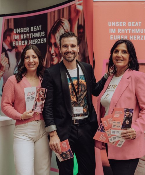 Drei Personen stehen lächelnd an einem Stand und halten Broschüren für eine hochzeits DJ Eventagentur in der Hand. Hinter ihnen sind Schilder mit deutschem Text und lebendigen Bildern zu sehen, die auf eine Messe in Österreich hinweisen, auf der für DJ Service und Eventplanung geworben wird.